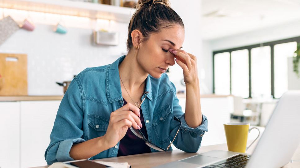 A tired employee sits in front of their laptop with a cup of coffee and their eyes closed.