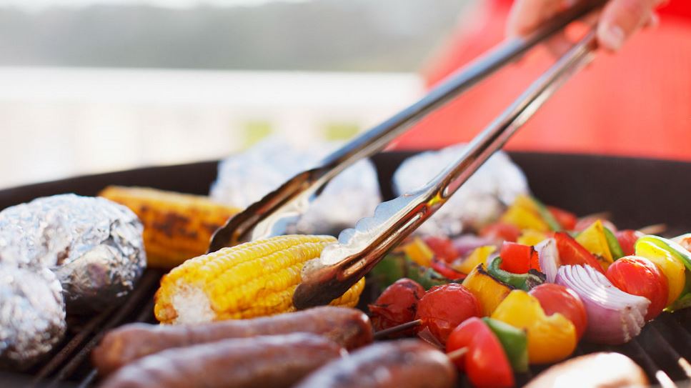 A pair of tongs grab hold of a corn cob on the grill, alongside other vegetables and meat.